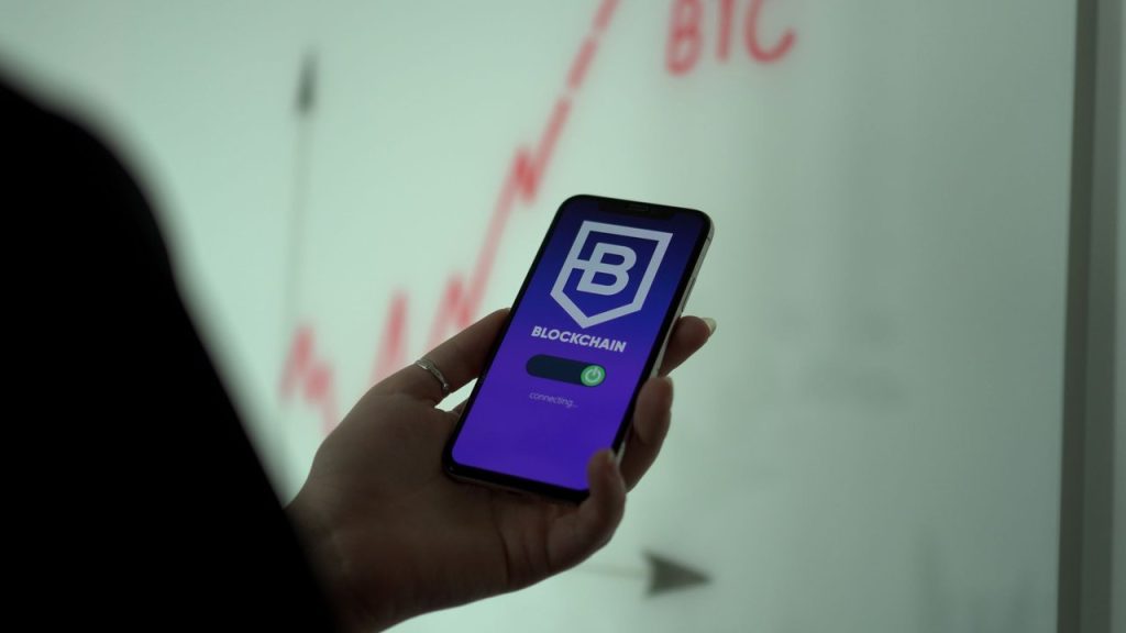 Mão segurando um smartphone com um aplicativo de blockchain, diante de um gráfico do mercado acionário.