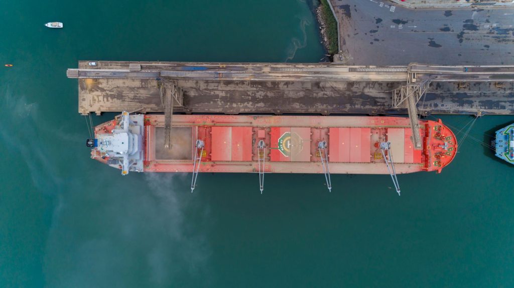 Vista aérea de um navio cargueiro atracado em um porto brasileiro, cercado por estruturas e áreas de movimentação marítima.